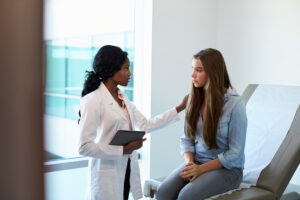 Female doctor speaking with unhappy teenage girl in exam room.